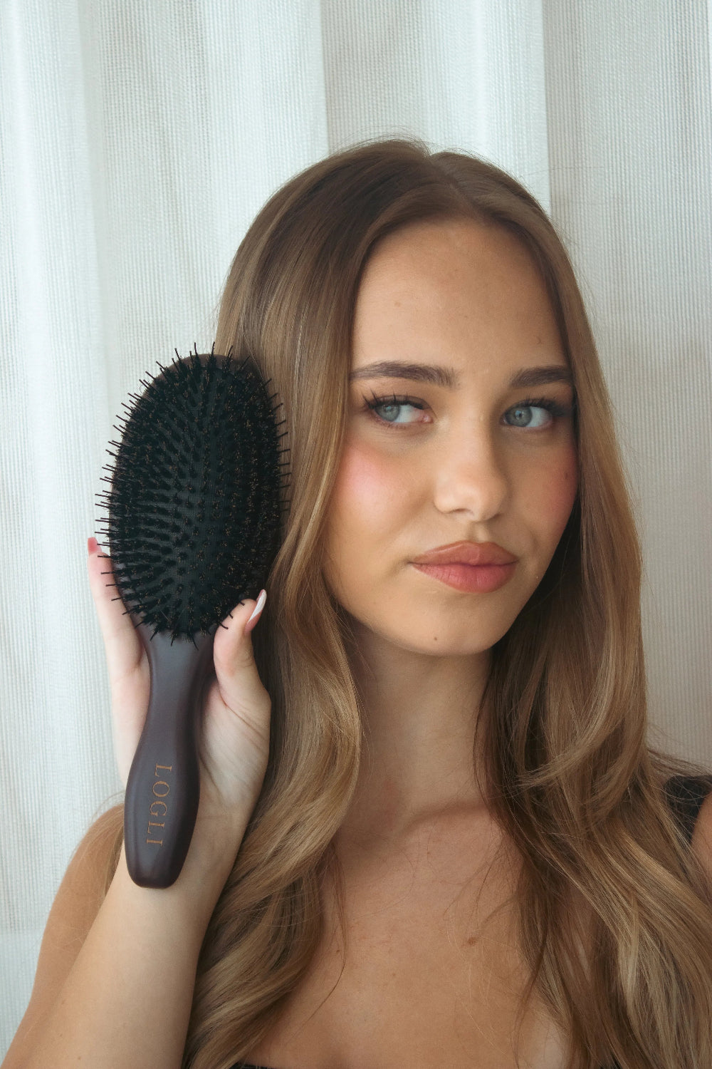 Woman holding a burgundy hairbrush with a white curtain background