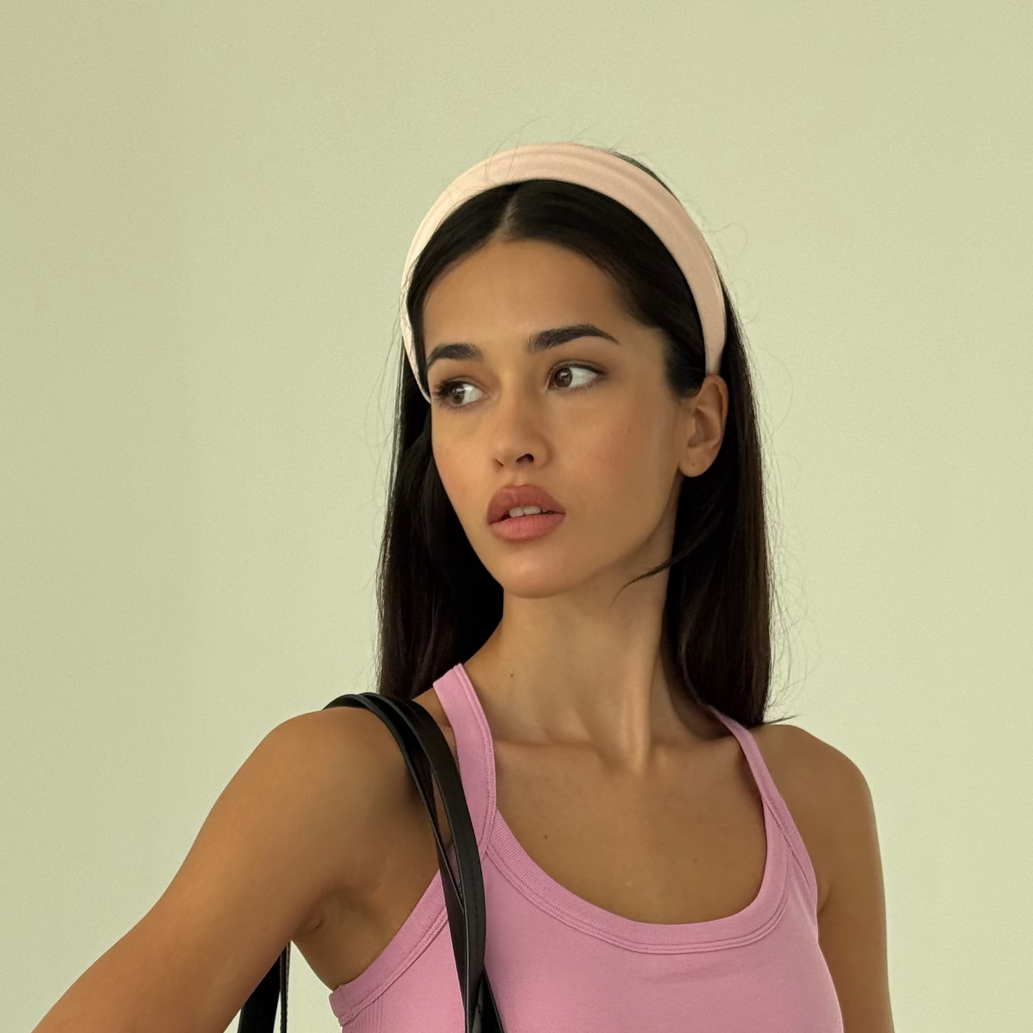 A woman wearing a pink tank top, red pants, and a pink headband carries a large black leather duffel bag with multiple compartments and handles, set against a solid pale green studio background.