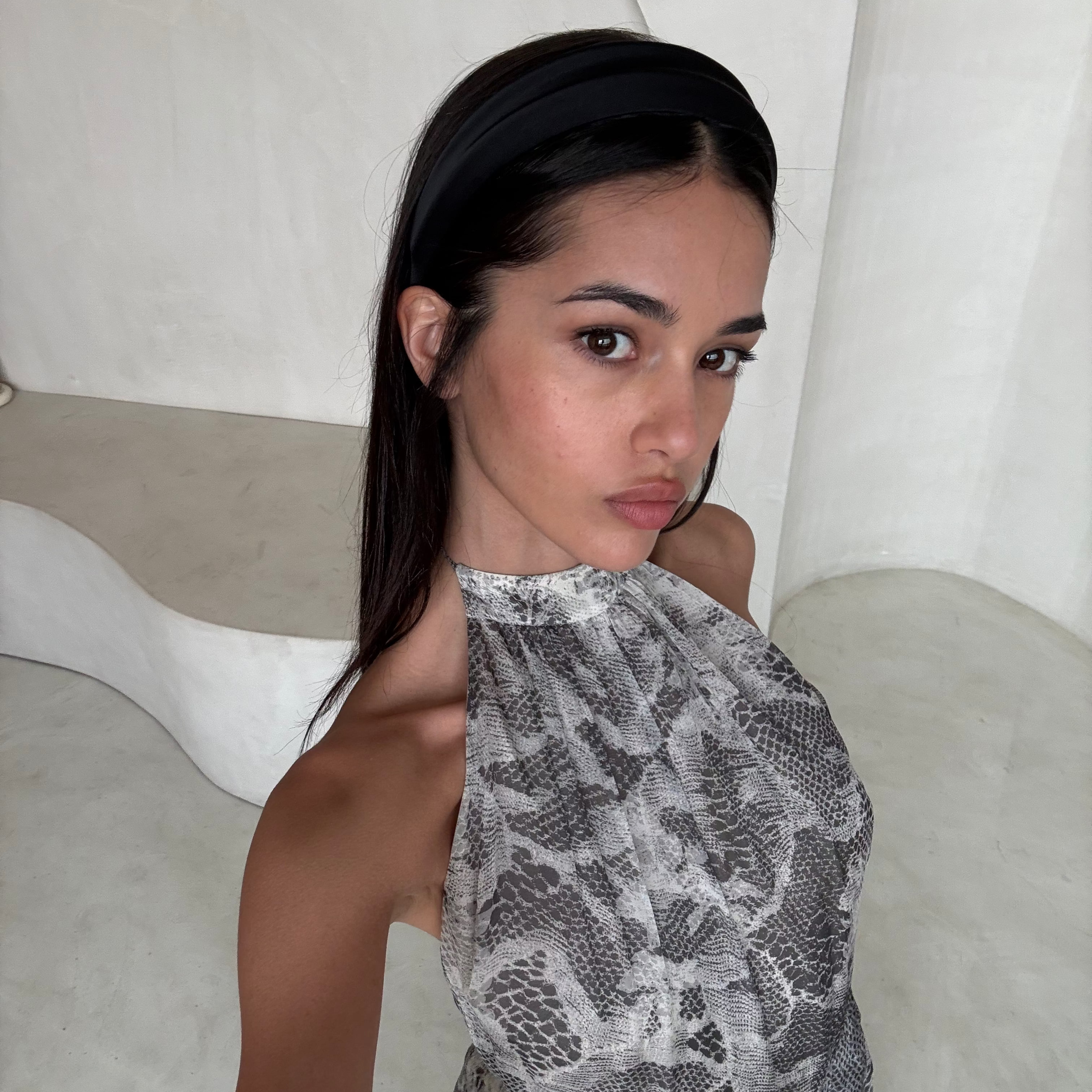A woman wearing a gray and white snakeskin print halter dress with a black headband, posing in a minimalist white room.