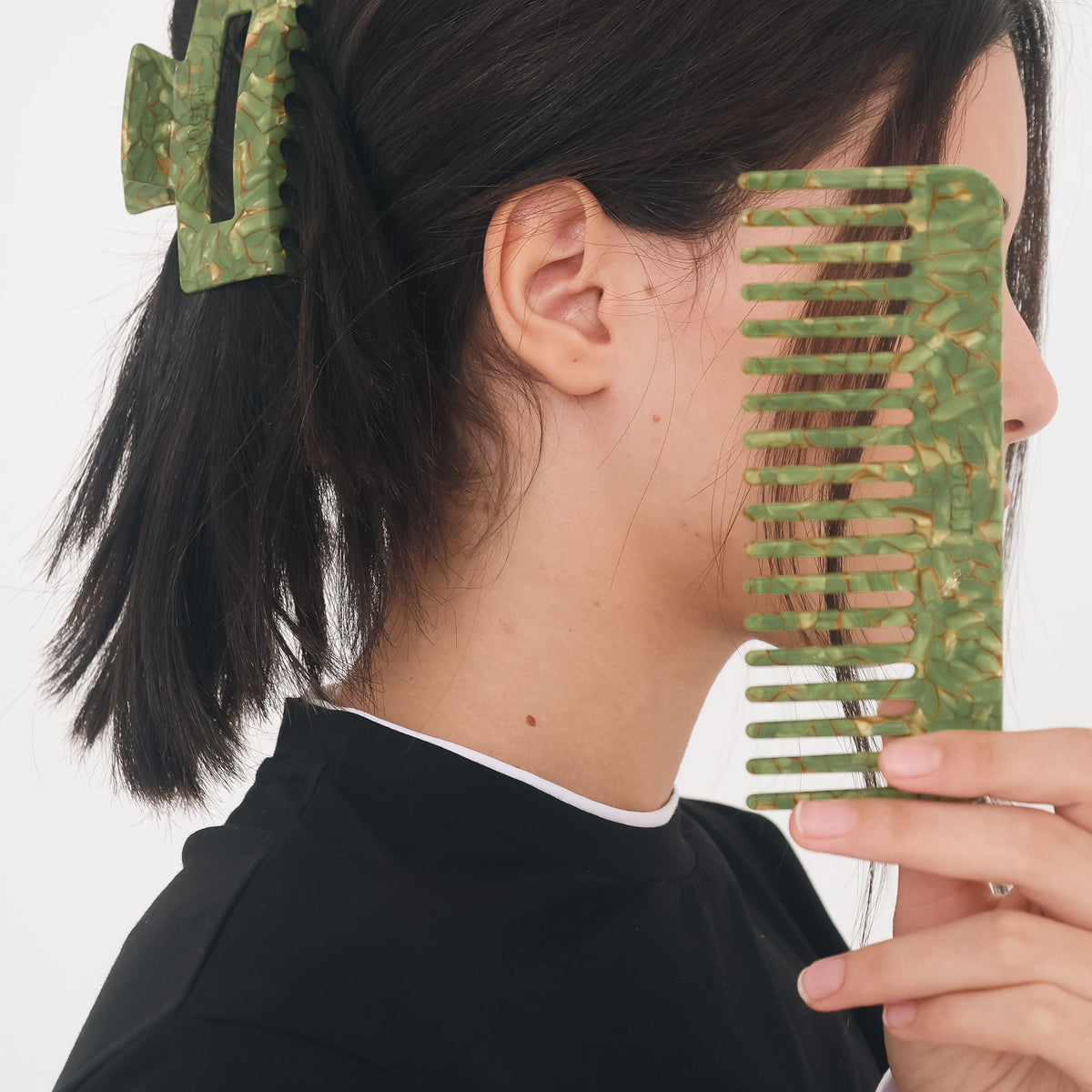 A person shown in side profile wearing a green camouflage hair claw clip and holding a matching green camouflage wide-tooth comb against their face, against a plain white background.