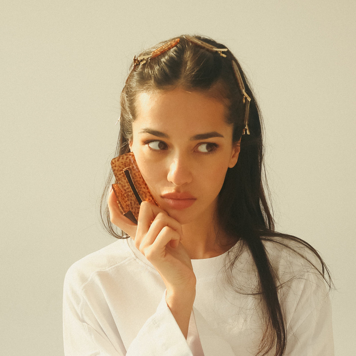 A woman with dark hair wearing a white long-sleeve top and a gold headband, holding a brown patterned claw to her ear against a plain light beige background.