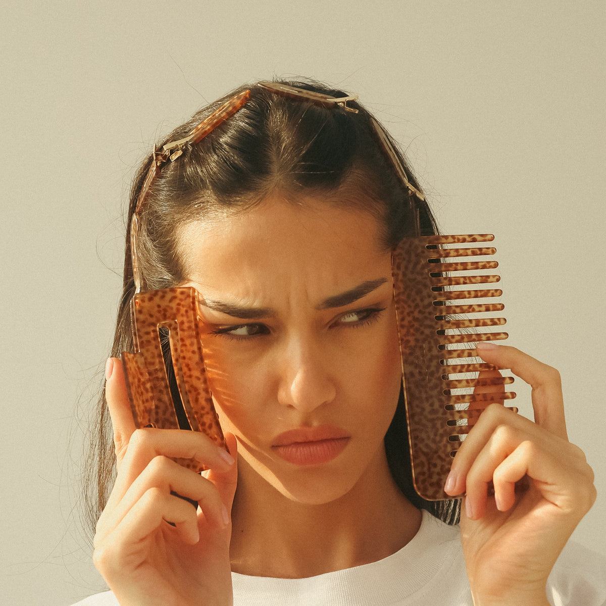 A woman holds two textured brown hair combs against her eyes, looking to the side with a skeptical or confused expression. She is wearing a white top against a plain, warm off-white background.