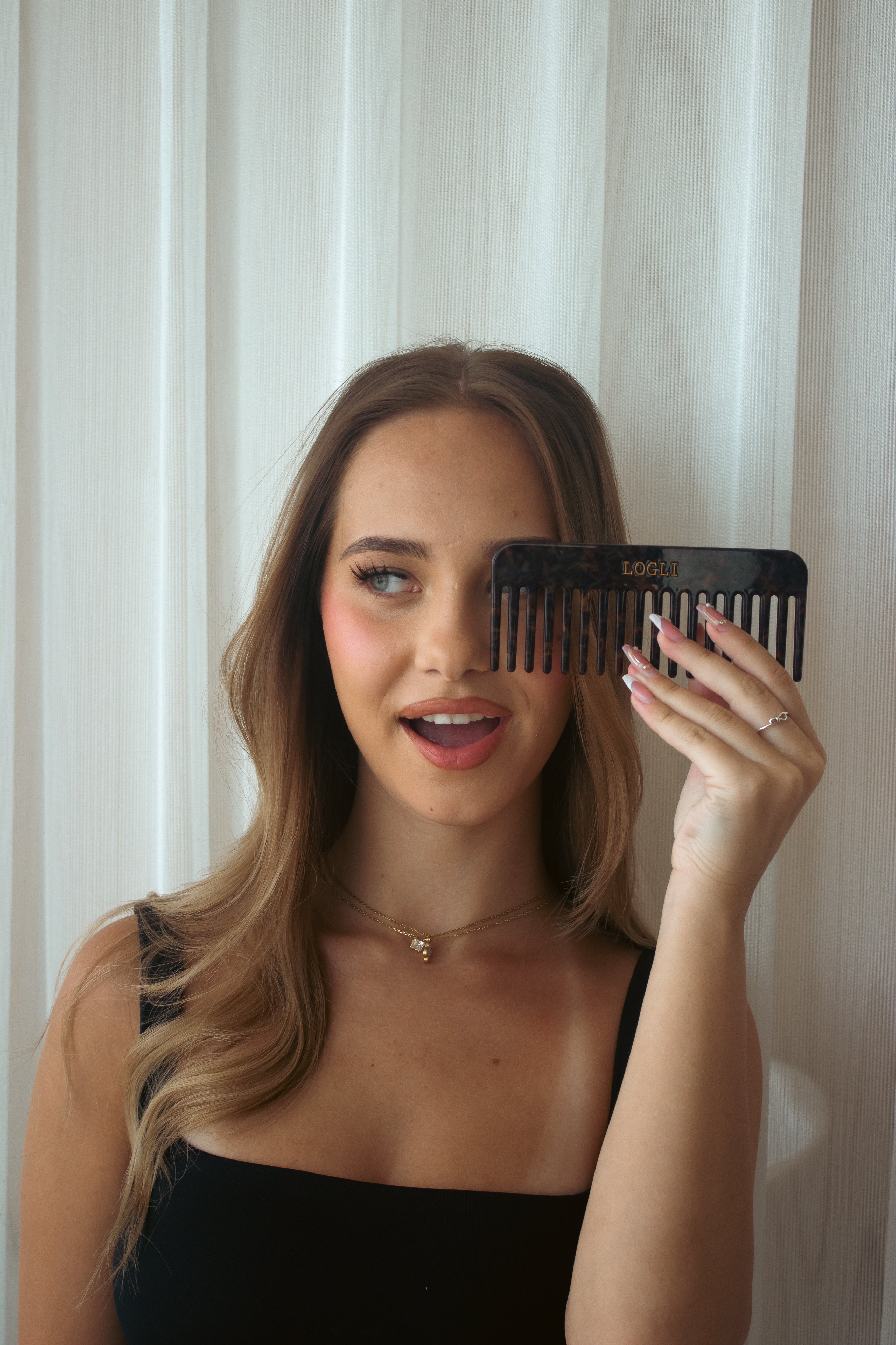 Woman holding a black comb in front of her face against a white curtain background