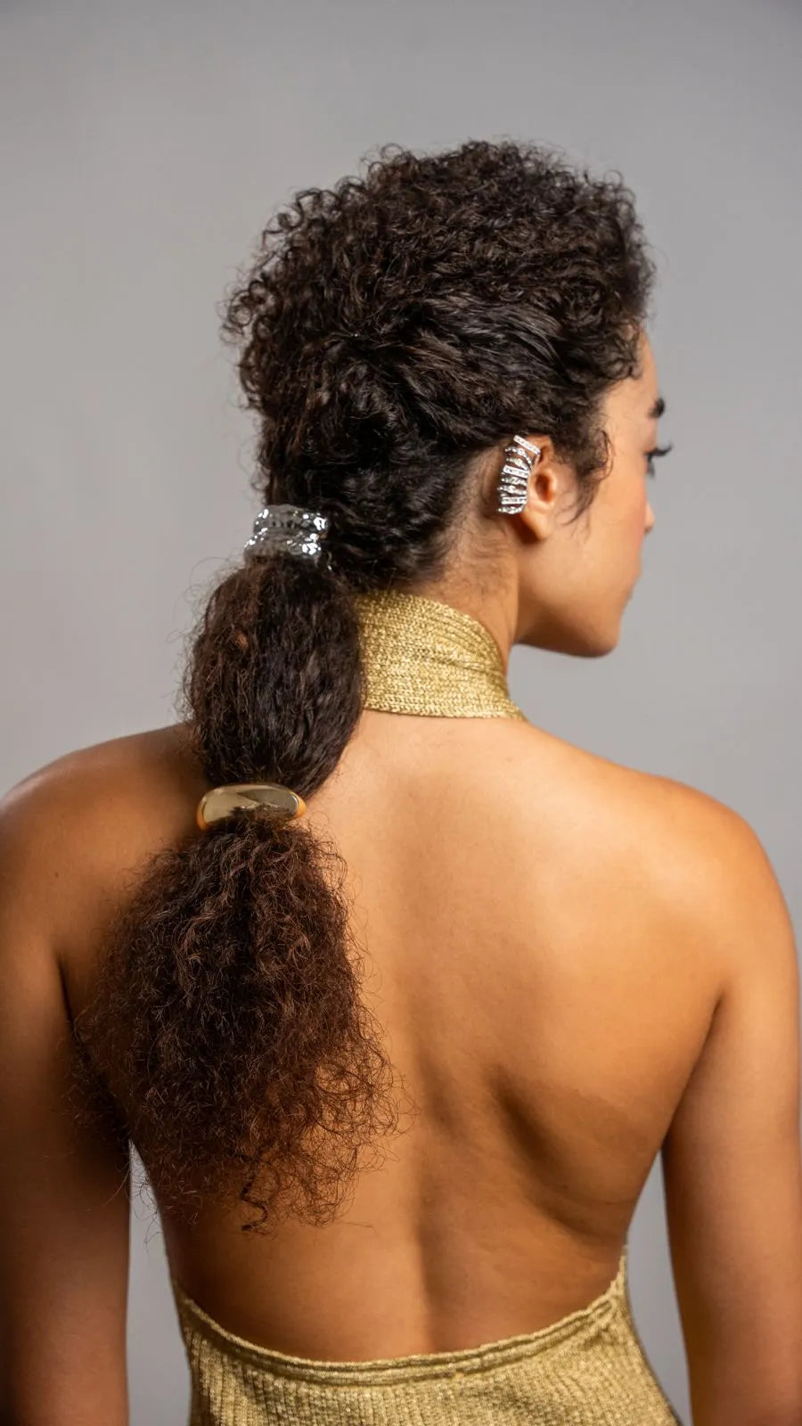 Woman with styled hair in a high ponytail wearing a gold garment against a gray background