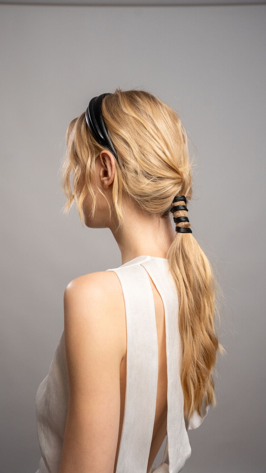 Woman with blonde hair styled in a ponytail with a black headband against a gray background